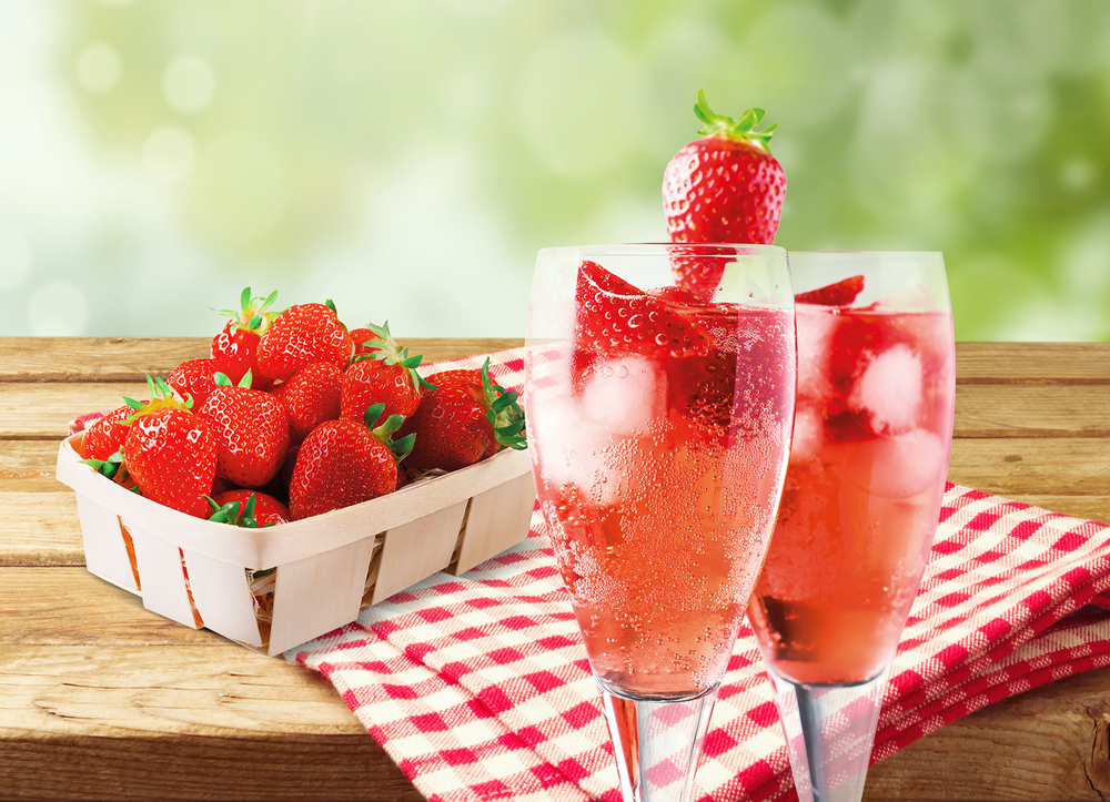 Frische Erdbeeren in einem Holzkorb neben zwei Gläsern spritzigem Erdbeergetränk mit Eiswürfeln, serviert auf einem rot-weiß karierten Tuch vor unscharfem grünem Hintergrund.