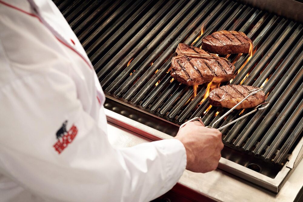 Blick über die Schulter eines Grillers, der eine Steakzange in der Hand hält und gerade ein Steak auf einem flammenden Grill wendet. 