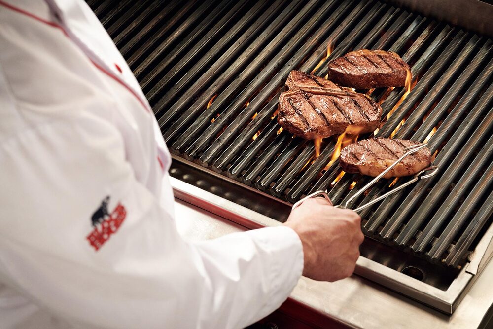 BLOCK HOUSE Steak auf dem Grill Blick über die Schulter eines Grillers, der eine Steakzange in der Hand hält und gerade ein Steak auf einem flammenden Grill wendet.