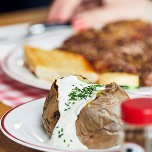 Essen Blick auf einen gedeckten Tisch im BLOCK HOUSE: Ein Glas Wasser, eine Baked Potato mit Sour Cream, Gewürze und im Hintergrund ein Steakteller