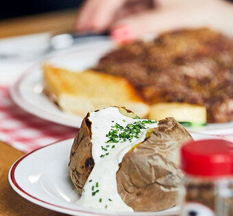 Blick auf einen gedeckten Tisch im BLOCK HOUSE: Ein Glas Wasser, eine Baked Potato mit Sour Cream, Gewürze und im Hintergrund ein Steakteller