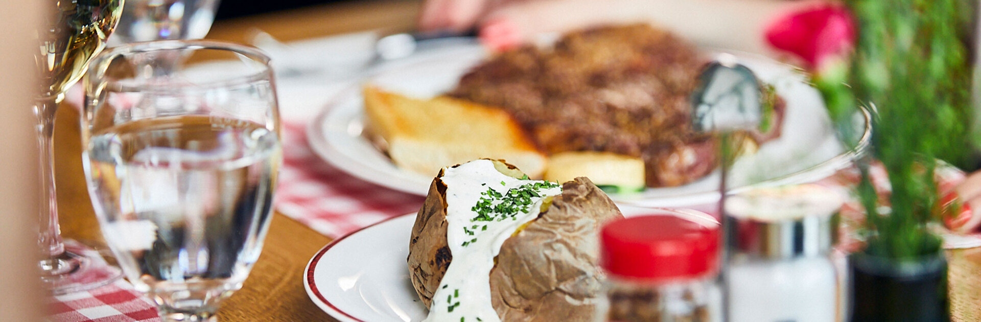 Blick auf einen gedeckten Tisch im BLOCK HOUSE: Ein Glas Wasser, eine Baked Potato mit Sour Cream, Gewürze und im Hintergrund ein Steakteller