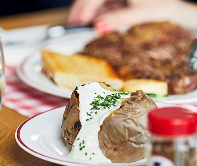 Blick auf einen gedeckten Tisch im BLOCK HOUSE: Ein Glas Wasser, eine Baked Potato mit Sour Cream, Gewürze und im Hintergrund ein Steakteller