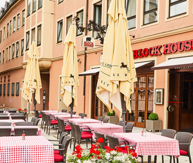 Außenansicht des BLOCK HOUSE Restaurants in Augsburg: eine große, hölzerne Fensterfront, davor viele Sitzgelegenheiten auf der Terrasse, die sich direkt auf dem Ulrichsplatz befindet.