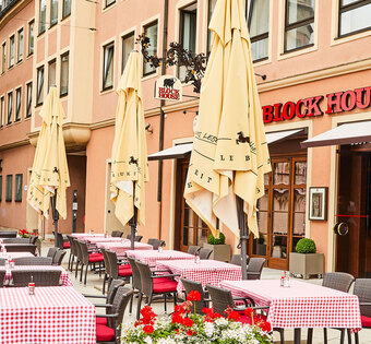 Außenansicht des BLOCK HOUSE Restaurants in Augsburg: eine große, hölzerne Fensterfront, davor viele Sitzgelegenheiten auf der Terrasse, die sich direkt auf dem Ulrichsplatz befindet.