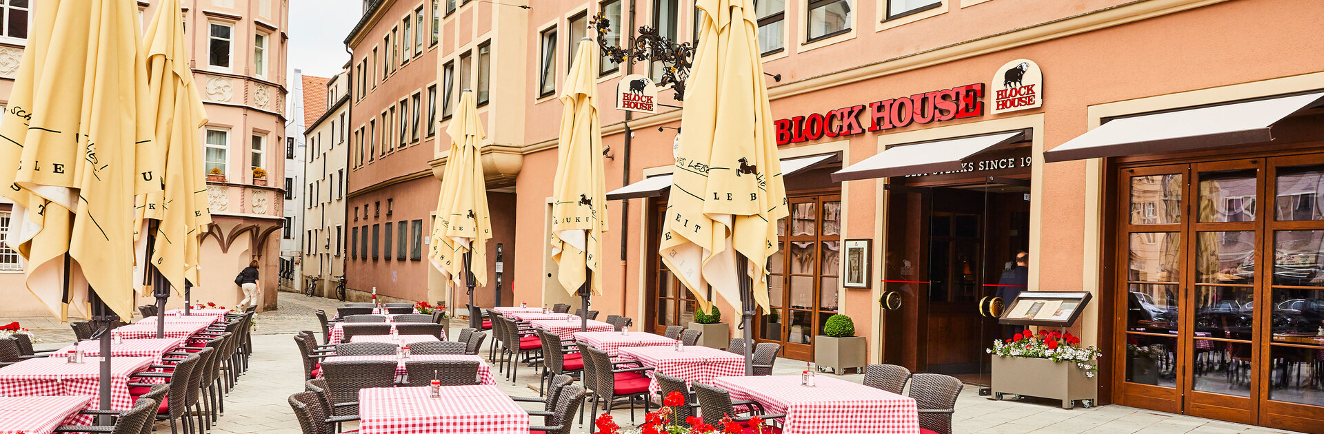 Außenansicht des BLOCK HOUSE Restaurants in Augsburg: eine große, hölzerne Fensterfront, davor viele Sitzgelegenheiten auf der Terrasse, die sich direkt auf dem Ulrichsplatz befindet.