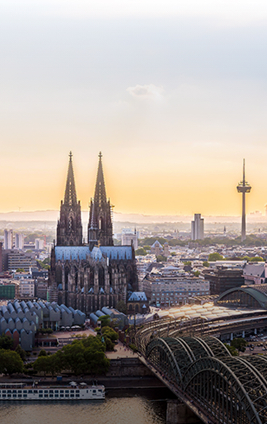 Blick über die Stadt Köln. Im Vordergrund ist die Hohenzollernbrücke zu sehen, dann der Kölner Dom. Im Hintergrund ist der Fernsehturm zu sehen. 