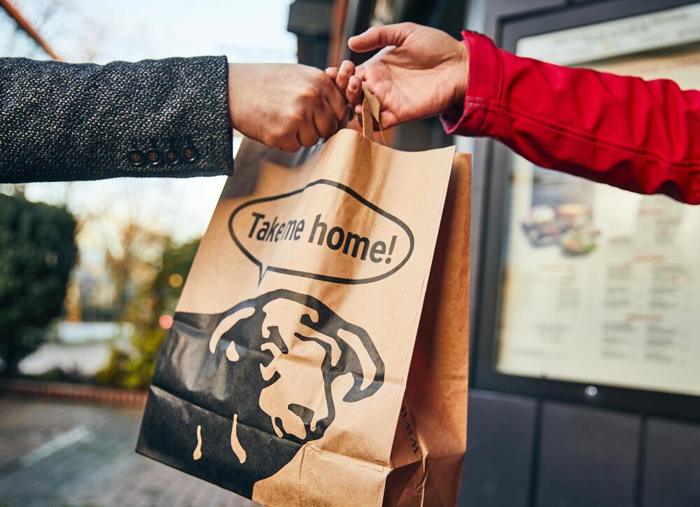 BLOCK HOUSE Mitarbeiter übergibt eine Takeaway Papiertüte mit der Aufschrift „Take me home!“ 