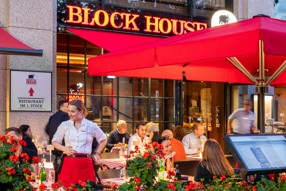 Terrasse des BLOCK HOUSE München Elisenhof mit Gästen und Servicepersonal unter roten Sonnenschirmen.