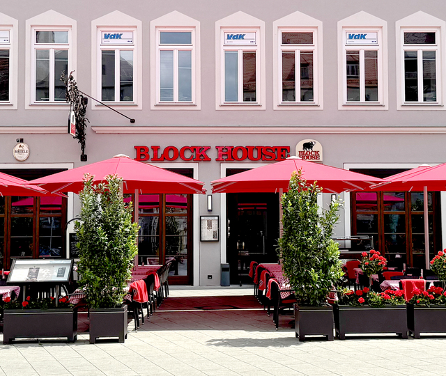 Außenansicht des BLOCK HOUSE Restaurants in Augsburg: eine große, hölzerne Fensterfront, davor viele Sitzgelegenheiten auf der Terrasse, die sich direkt auf dem Ulrichsplatz befindet.