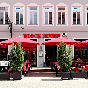 Außenansicht des BLOCK HOUSE Restaurants in Augsburg: eine große, hölzerne Fensterfront, davor viele Sitzgelegenheiten auf der Terrasse, die sich direkt auf dem Ulrichsplatz befindet.