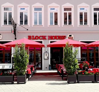 Außenansicht des BLOCK HOUSE Restaurants in Augsburg: eine große, hölzerne Fensterfront, davor viele Sitzgelegenheiten auf der Terrasse, die sich direkt auf dem Ulrichsplatz befindet.