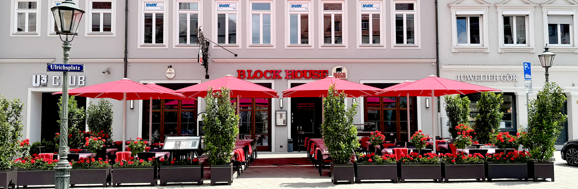 Außenansicht des BLOCK HOUSE Restaurants in Augsburg: eine große, hölzerne Fensterfront, davor viele Sitzgelegenheiten auf der Terrasse, die sich direkt auf dem Ulrichsplatz befindet.