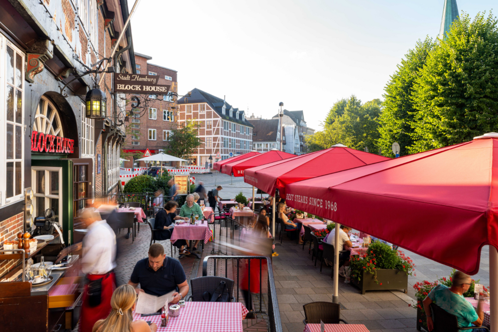Terrasse des BLOCK HOUSE Hamburg Bergedorf mit roten Sonnenschirmen, gedeckten Tischen und Gästen vor dem historischen Fachwerkgebäude.