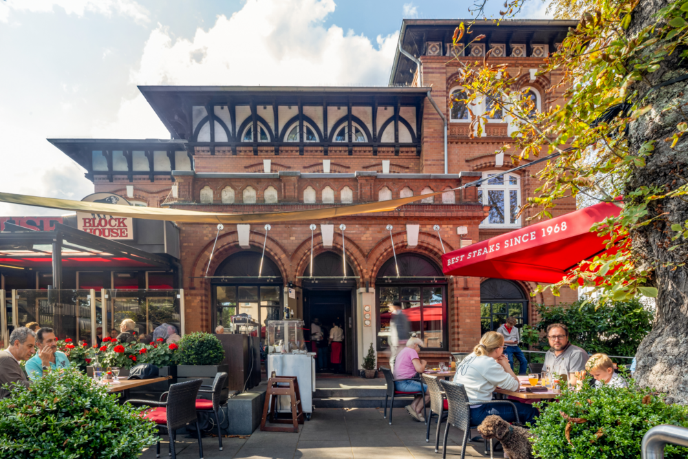 Eingangsbereich des BLOCK HOUSE Hamburg Othmarschen mit Terrasse, Gästen und roten Sonnenschirmen vor dem Steakhouse