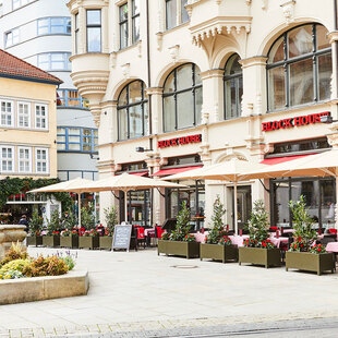 Außenansicht des BLOCK HOUSE Restaurants in Erfurt in einem denkmalgeschützten Gebäude . Links ist der alte Angerbrunnen zu sehen. Die Terrasse des Restaurants ist von Pflanzkübeln umsäumt, die Sonnenschirme sind aufgespannt. Das Restaurant hat große Fensterfronten, die den direkten Blick auf den alten Angerbrunnen ermöglichen.  