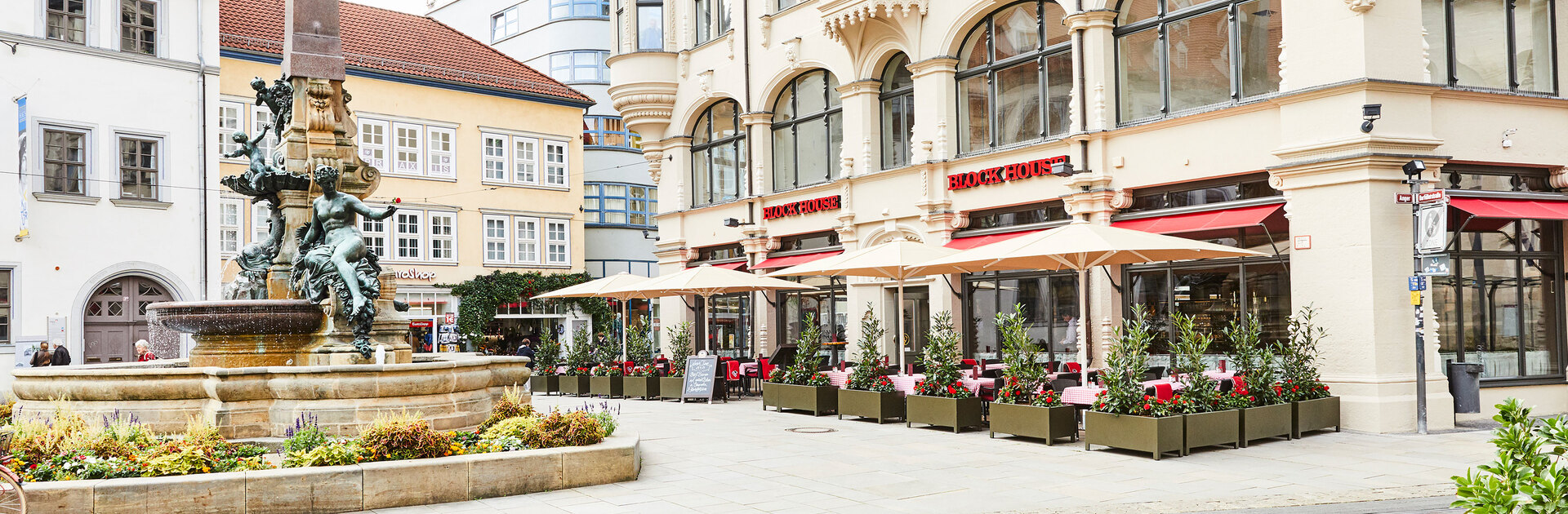 Außenansicht des BLOCK HOUSE Restaurants in Erfurt in einem denkmalgeschützten Gebäude . Links ist der alte Angerbrunnen zu sehen. Die Terrasse des Restaurants ist von Pflanzkübeln umsäumt, die Sonnenschirme sind aufgespannt. Das Restaurant hat große Fensterfronten, die den direkten Blick auf den alten Angerbrunnen ermöglichen.  