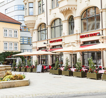 Außenansicht des BLOCK HOUSE Restaurants in Erfurt in einem denkmalgeschützten Gebäude . Links ist der alte Angerbrunnen zu sehen. Die Terrasse des Restaurants ist von Pflanzkübeln umsäumt, die Sonnenschirme sind aufgespannt. Das Restaurant hat große Fensterfronten, die den direkten Blick auf den alten Angerbrunnen ermöglichen.  