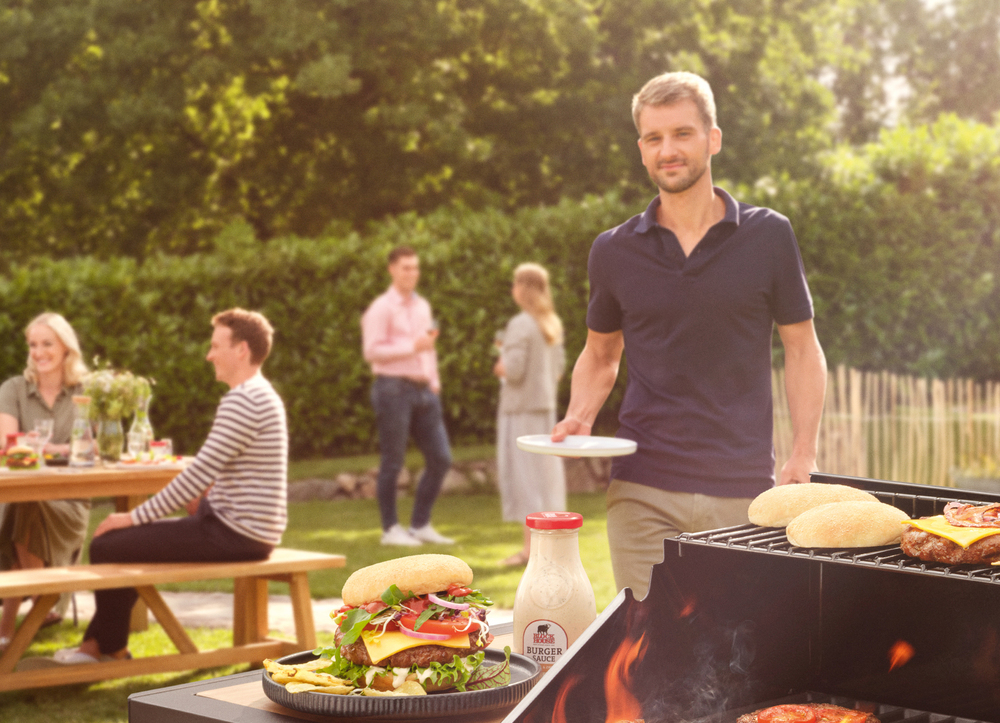 Im Vordergrund ein Grill mit BLOCK HOUSE Burgern und Sauce; links sitzen mehrere Personen an einem Tisch, im Hintergrund unterhalten sich ein Mann und eine Frau; ein weiterer Mann kommt mit seinem Teller auf den Grill zu