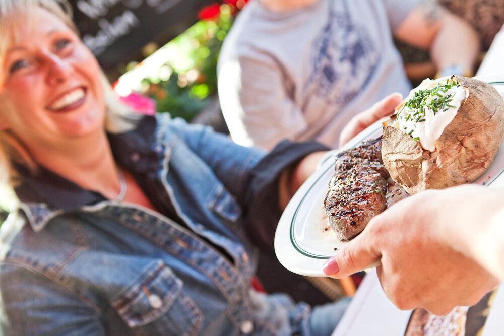 Servicekraft serviert Teller mit Steak und BLOCK HOUSE Baked Potato Eine Servicekraft serviert einer lächelnden Gästin einen Teller mit Steak und BLOCK HOUSE Baked Potato