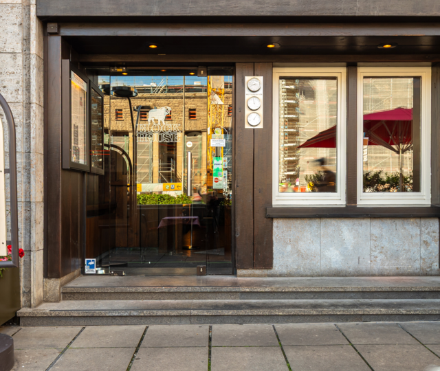 Eingang des BLOCK HOUSE Stuttgart Arnulf-Klett-Platz mit Menütafel vor dem Steakhouse und Gästen an Tischen mit rot-weiß karierten Tischdecken