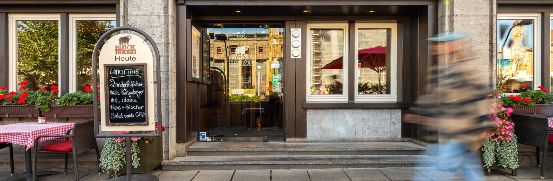 Eingang des BLOCK HOUSE Stuttgart Arnulf-Klett-Platz mit Menütafel vor dem Steakhouse und Gästen an Tischen mit rot-weiß karierten Tischdecken
