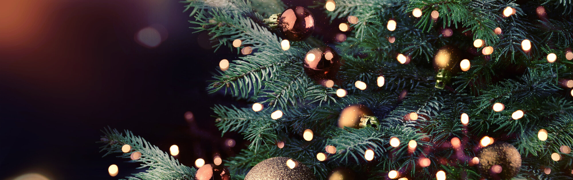 Nahaufnahme eines festlich geschmückten Weihnachtsbaums mit glänzenden goldenen und bronzefarbenen Kugeln sowie warm leuchtenden Lichterketten. Im Hintergrund erzeugen verschwommene Lichter eine gemütliche, festliche Stimmung.