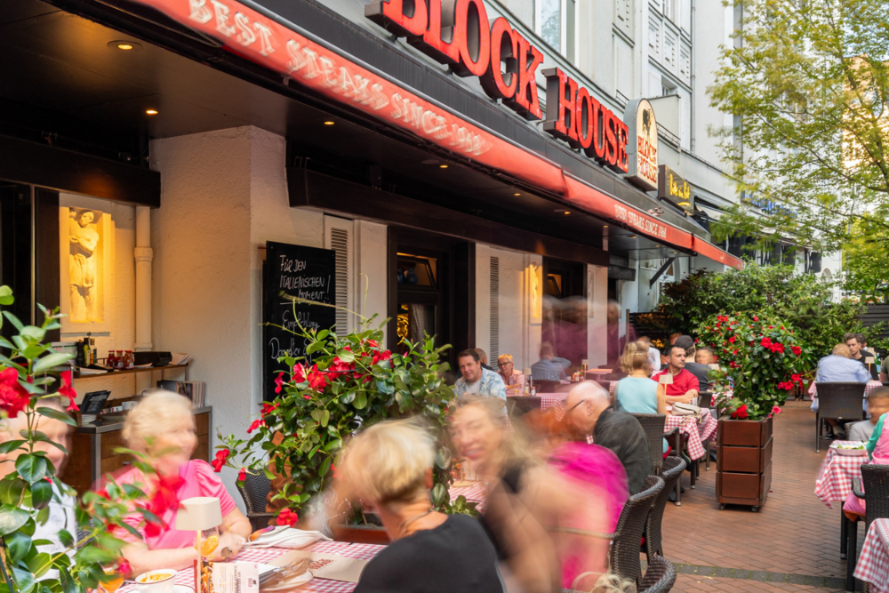 Terrasse des BLOCK HOUSE Berlin Theodor-Heuss-Platz mit Gästen an Tischen mit rot-weiß karierten Tischdecken vor dem Restaurant