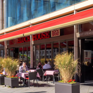 Außenansicht des BLOCK HOUSE Frankfurt Europa-Allee mit ausgefahrener Markise und Gästen auf der Terrasse bei gutem Wetter