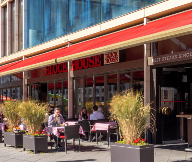 Außenansicht des BLOCK HOUSE Frankfurt Europa-Allee mit ausgefahrener Markise und Gästen auf der Terrasse bei gutem Wetter