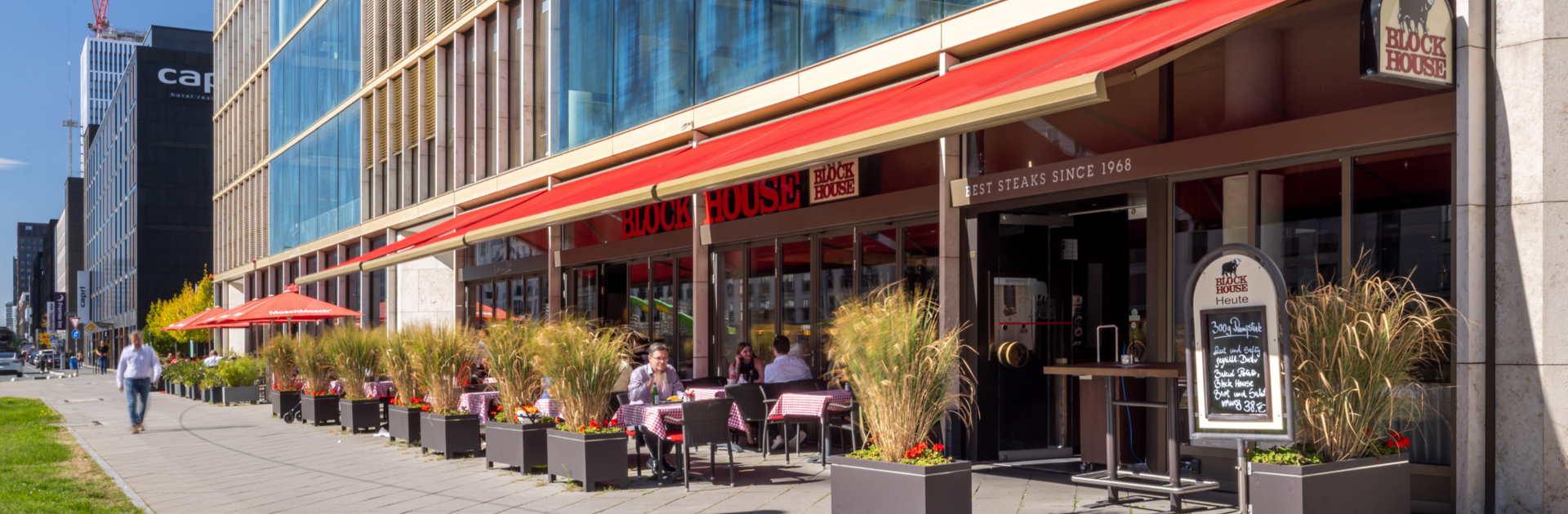 Außenansicht des BLOCK HOUSE Frankfurt Europa-Allee mit ausgefahrener Markise und Gästen auf der Terrasse bei gutem Wetter