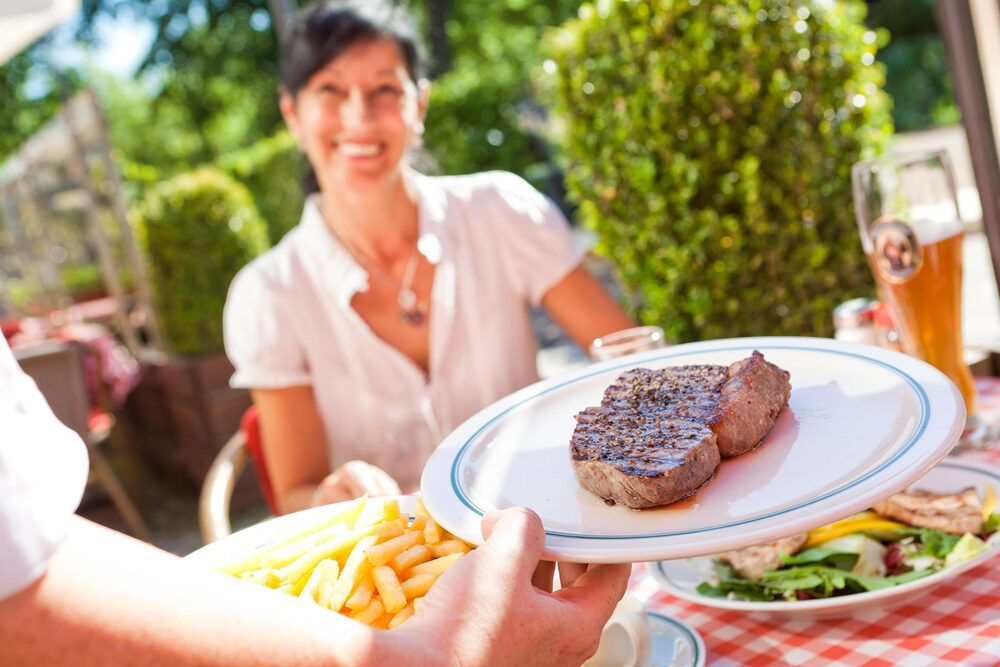 Man sieht im Vordergrund den Arm einer Kellnerin. Sie hält einen Teller mit einem Steak, aus dem Saft austritt. Zusätzlich hält sie einen Teller mit Pommes frites. Im Hintergrund sitzt eine lächelnde Frau, die vor sich auf dem Tisch bereits einen Teller Salat stehen hat. Hinter ihr ist die Terrasse des BLOCK HOUSE Restaurants in Ahrensburg zu sehen. 