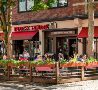 BLOCK HOUSE Restaurant in Kiel Außenansicht des BLOCK HOUSE Kiel mit Terrasse, roten Markisen und Gästen vor dem Restaurant.