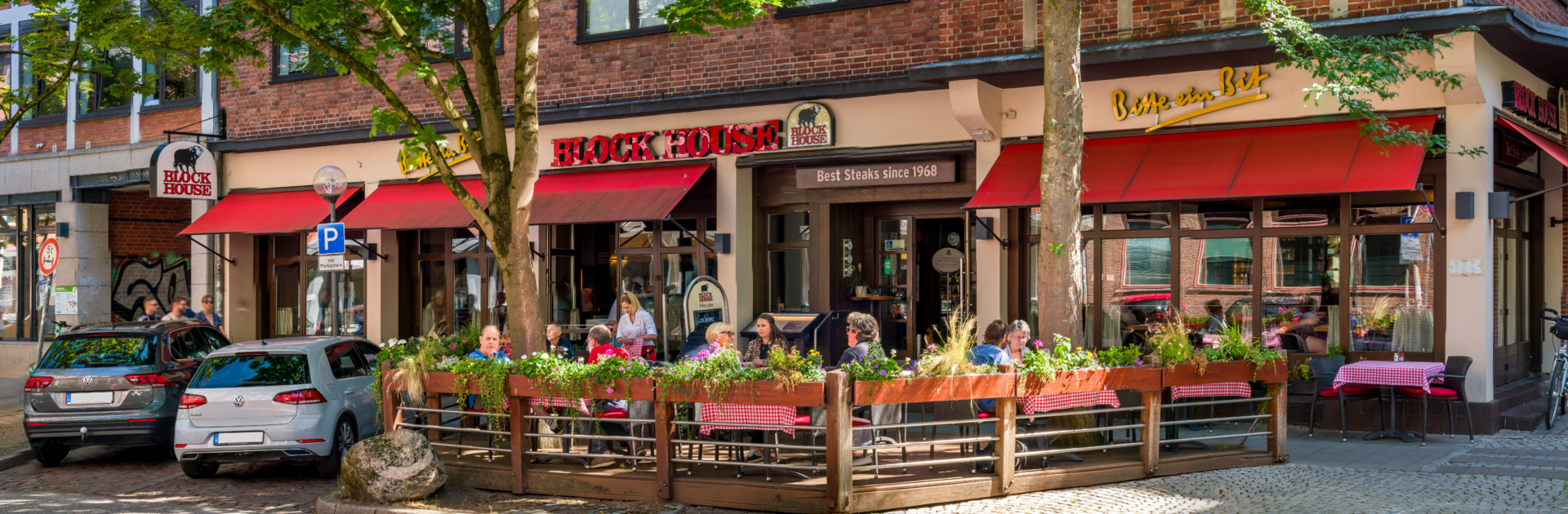 BLOCK HOUSE Restaurant in Kiel Außenansicht des BLOCK HOUSE Kiel mit Terrasse, roten Markisen und Gästen vor dem Restaurant.