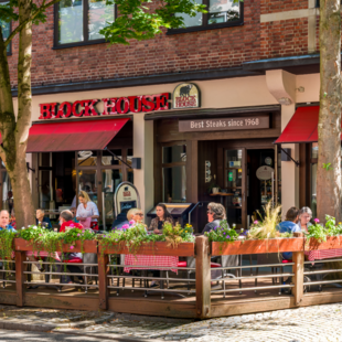 BLOCK HOUSE Restaurant in Kiel Außenansicht des BLOCK HOUSE Kiel mit Terrasse, roten Markisen und Gästen vor dem Restaurant.