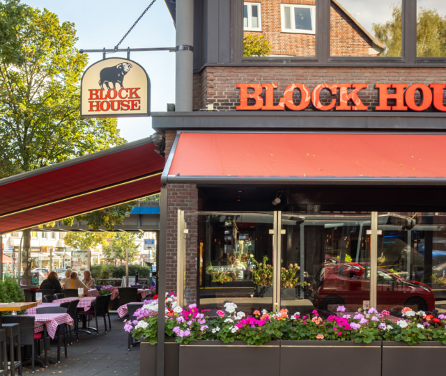 Außenansicht des BLOCK HOUSE Hamburg Barmbek mit Terrasse, roten Markisen und Sitzplätzen vor dem Steakhouse