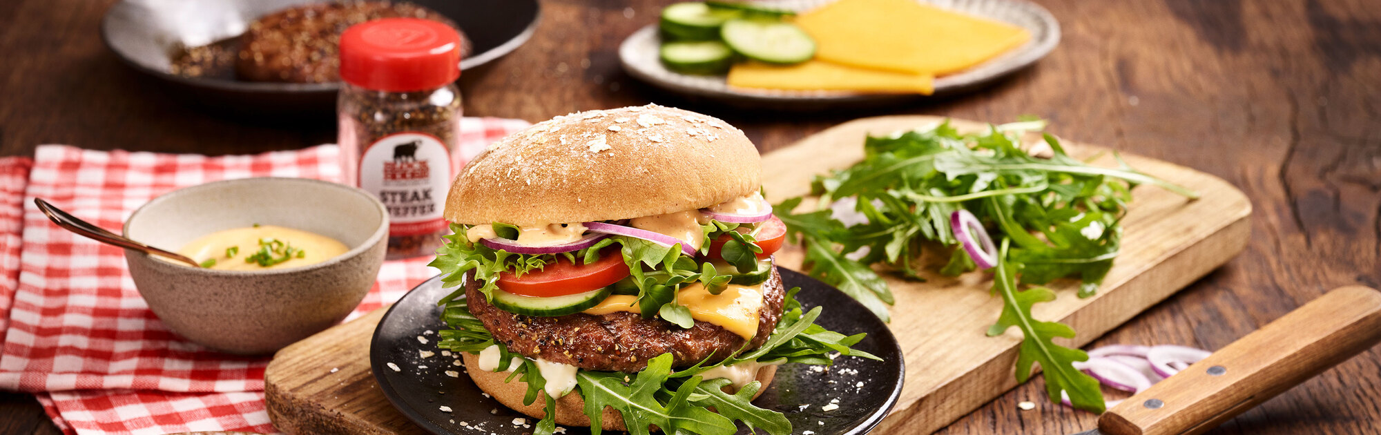 BLOCK HOUSE Burger mit Fleischpatty, Rucola, Tomate, Salat , Zwiebeln und Sauce auf einem Holzbrett mit verwendeten Zutaten im Hintergrund