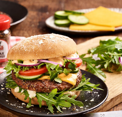 BLOCK HOUSE Burger mit Fleischpatty, Rucola, Tomate, Salat , Zwiebeln und Sauce auf einem Holzbrett mit verwendeten Zutaten im Hintergrund