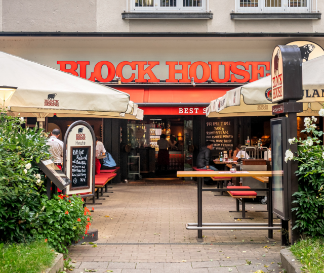 BLOCK HOUSE München Leopoldstraße – Steakhouse mit Terrasse Eingang und Terrasse des BLOCK HOUSE München Leopoldstraße mit Sonnenschirmen und Gästen vor dem Steakhouse